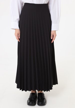 Falda plisada negra de talle alto, combinada con camisa blanca de rayas finas de manga larga y mocasines negros brillantes, visible desde la cintura hasta los pies.