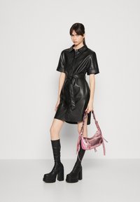 Zwarte neplederen hemdjurk met korte mouwen, een getailleerde taille en een knoopsluiting aan de voorkant. Gepaarde met kniehoge zwarte laarzen en een roze metallic handtas.