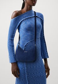 Niebieska dżinsowa torba na ramię o zaokrąglonym kształcie i srebrnych akcentach, z logo Levi's. Połączona z dopasowaną, prążkowaną niebieską sukienką.