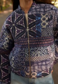 Vrouw in een ritsjack met blauwe, paarse en beige patchworkpatronen en lichtblauwe jeans.