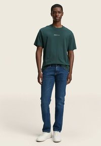 Tamnozelena pamučna majica s okruglim izrezom i kratkim rukavima, uparena s plavim trapericama i bijelim tenisicama, stoji na neutralnoj pozadini.