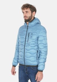 Giacca imbottita azzurro chiaro con cappuccio, caratterizzata da un motivo a onde, chiusura frontale con zip e due tasche laterali. Texture morbida e orlo regolabile.