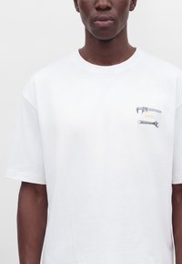 Personne portant un t-shirt blanc avec de petits graphiques de clé et clé à molette et du texte jaune sur la partie gauche de la poitrine.