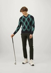 Marinblå tröja med tealfärgad argyle-mönster, V-halsdesign; kombinerad med svarta byxor och vita sneakers; håller en golfklubba.