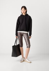 Zwarte hoodie, leopardprint fietsbroek, zilveren sneakers en een zwarte totebag. Model toont een ontspannen, sportieve uitstraling.