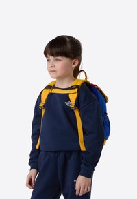 Zils un dzeltens mugursoma ar regulējamām lencēm un ārēju kabatu. Bērns, tērpies tumši zilos džemperī, stāv pret vienkāršu fona sienu.