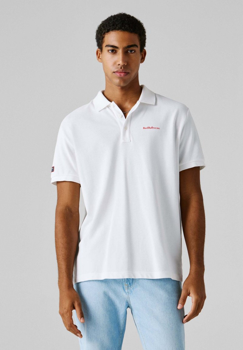 Νέος άνδρας που φοράει λευκό πόλο μπλουζάκι Red Bull Racing και ανοιχτό μπλε τζιν, στέκεται μπροστά σε απλό γκρι φόντο.