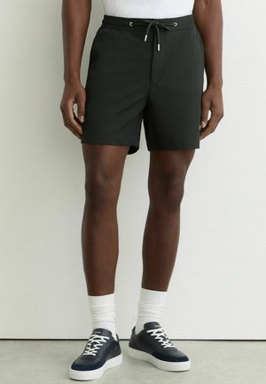 Mężczyzna w czarnych szortach ze sznurkiem, białych skarpetkach za kostkę i czarnych niskich trampkach, stojący na tle gładkiej białej ściany.