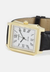 Montre rectangulaire en or avec un cadran blanc, des chiffres romains noirs et des aiguilles argentées. Bracelet en cuir noir texturé avec des détails de couture. Résistante à l'eau.