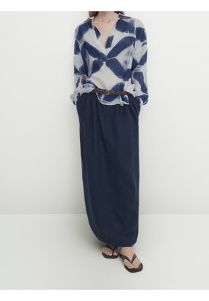 Blouse légère à motifs blancs et marins, associée à une jupe ample bleu foncé et des tongs noires. Une ceinture tressée marron souligne la taille.