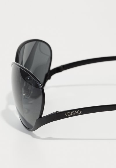 Ochelari de soare mari, negri, cu lentile întunecate și un cadru elegant, lucios. Prezintă un logo argintiu "VERSACE" pe bradă. Textură fină pe toată suprafața.