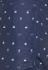 Shorts de bain navy avec un imprimé nautique blanc répété de têtes de mort, voiliers, pagaies croisées et nageoires, avec une texture lisse et des coutures cousues.