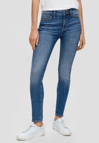 Jeans attillati in denim blu medio con cuciture visibili, tasche frontali e un design aderente, abbinati a scarpe sportive bianche.