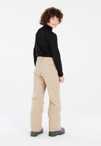 Pantaloni da sci beige con una texture liscia, caratterizzati da un vita alta e dettagli con tasche posteriori, abbinati a un top nero aderente a maniche lunghe e calzature nere.