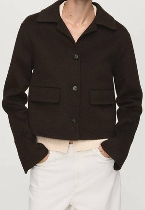 Veste noire courte avec col, trois boutons, poches à rabat, portée sur un top beige côtelé et un pantalon blanc, mains dans les poches.