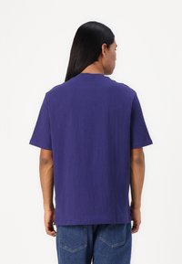 Camisa corta de manga morada con un tejido texturizado. Diseño de cuello redondo, corte relajado, dobladillo recto, combinada con jeans de mezclilla azules.