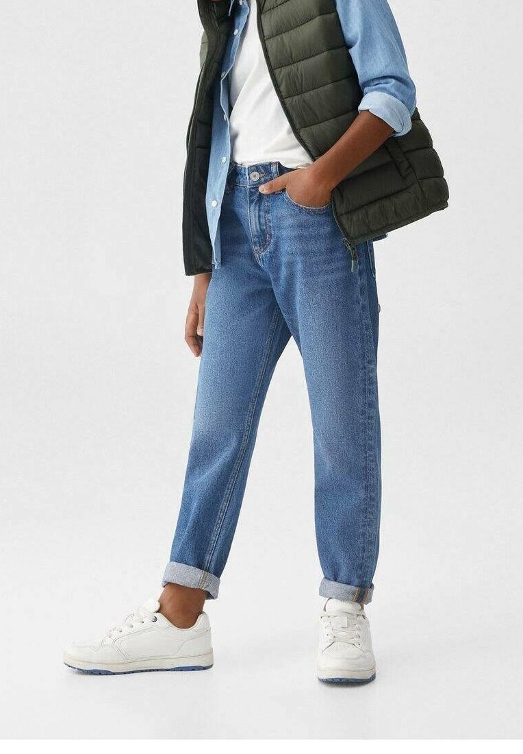 Personne portant un jean bleu clair retroussé, des baskets blanches, un t-shirt blanc, une chemise en denim bleue et une veste matelassée noire, debout avec une main dans la poche.