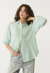 Mint zelena, dolgo rokav shirt iz lahkega materiala. Ima zapenjanje spredaj, sproščen kroj in zavihek rokavov. V kombinaciji z sivo jeans hlačami.