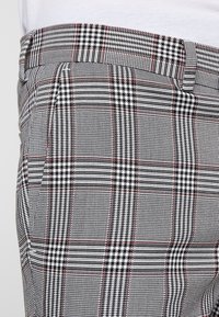 Szare i czarne spodnie w kratę z teksturowanej tkaniny, dopasowany krój oraz czerwone akcenty w formie przeszyć na kieszeniach i w pasie.