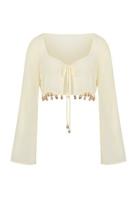 Blouse cropped à manches longues couleur crème avec un nœud à l'avant et des charms en coquillage dorés décoratifs le long de l'ourlet.