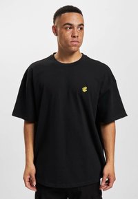 Černé oversized tričko vyrobené z bavlny, s krátkým rukávem a kulatým výstřihem. Malé žluté logo na levé straně hrudi.