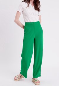 Femme portant un pantalon large vert, un haut en tricot texturé à manches courtes blanc, et des sandales à plateforme dorées avec des brides croisées.