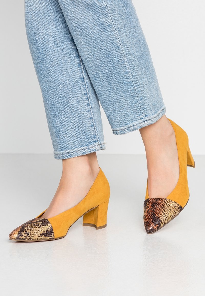 Scarpe con tacco alto in camoscio giallo, con punta affusolata e un accento a stampa serpente in contrasto nella parte anteriore, indossate con jeans azzurri chiari.
