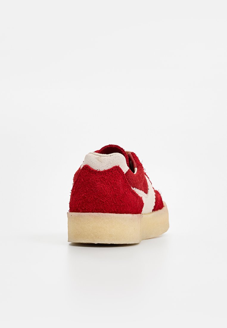 Baskets en suède rouge avec une accent en tissu beige, semelle en caoutchouc plate et logo blanc contrasté sur le côté. Vue de dos présentée.