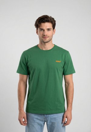 Hombre vistiendo una camiseta verde de manga corta con el texto "HERO" en el pecho y vaqueros azul claro, posando frente a un fondo liso gris claro.