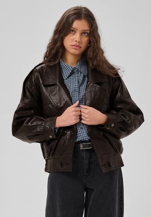 Junge Frau mit langen, lockigen Haaren trägt eine dunkelbraune Lederjacke über einem blau karierten Hemd und schwarzen Hosen vor einem einfarbigen Hintergrund.