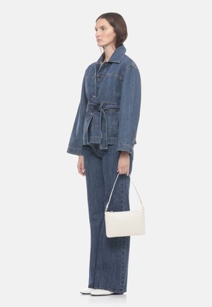 Donna che indossa una giacca di jeans blu con cintura da annodare e jeans a gamba larga abbinati, tenendo una piccola borsa bianca con tracolla corta.