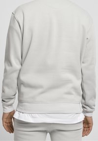 Felpa grigio chiaro con polsini e orlo a coste. Presenta un collo tondo e un orlo di una camicia bianca a strati visibile sotto. Tessuto morbido e testurizzato.