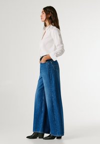 High-waisted wide-leg denim jeans in medium blue, featuring a classic five-pocket design and subtle stitching. Paired with a white button-up shirt.