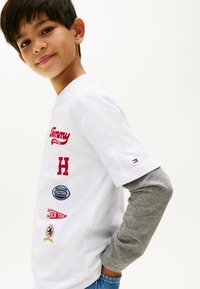 T-shirt bianco in cotone con loghi rossi e navy, tra cui "Tommy" e "H". Maglietta a maniche lunghe grigia sotto, con pantaloni corti in denim blu visibili in vita.