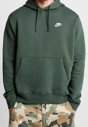 Mężczyzna w ciemnozielonej bluzie Nike z kapturem i kieszenią kangurka oraz w spodniach moro, stojący z rękami w kieszeniach.