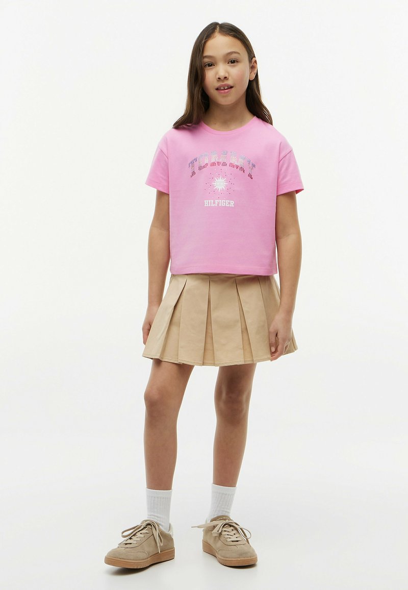 Jeune fille portant un t-shirt rose Tommy Hilfiger, une jupe plissée beige, des chaussettes blanches et des baskets beiges, debout devant un fond blanc uni.