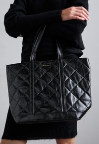 Geantă tote din piele matlasată neagră, cu mânere duble, formă rectangulară și detalii de cusătură vizibile. Prezintă un accent cu logo auriu.