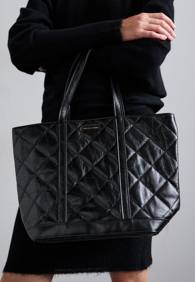 Sac fourre-tout en cuir matelassé noir avec poignées duales, de forme rectangulaire, et des détails de couture visibles. Présente un accent logo doré.