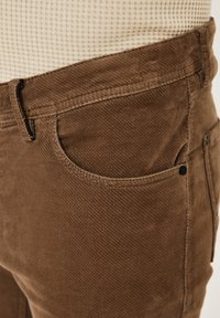 Brown corduroy pants with a textured surface, featuring a front pocket, zipper, and a subtle woven pattern.