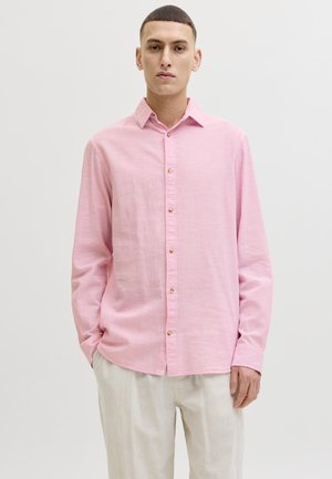 Noor mees seisab käed taskutes, seljas hele roosa nööbiga särk ja helevalged püksid, taust on ühtlane.