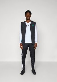 Μαύρο αθλητικό γιλέκο πάνω από άσπρο πουκάμισο με μακριά μανίκια, συνδυασμένο με μαύρο παντελόνι με λευκές ρίγες. Το ύφασμα φαίνεται ελαφρύ και διαπνέον.