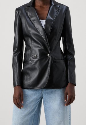 Osoba nosząca dopasowaną czarną skórzaną marynarkę dwurzędową na białej bluzce i jasnoniebieskich dżinsach, stojąca na jednolitym tle.