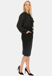 Czarna bluzka w kropki z falbaną z przodu, zestawiona z dopasowaną czarną spódnicą ołówkową. Modelka nosi czarne, szpiczaste szpilki. Gładka faktura materiału.