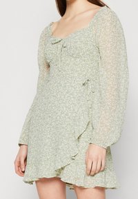 Ljusgrön blommig klänning med långa genomskinliga puffärmar, hjärtformad halsringning, frontdetaljer med knytning och volangkant i asymmetrisk stil, bärs av en person.