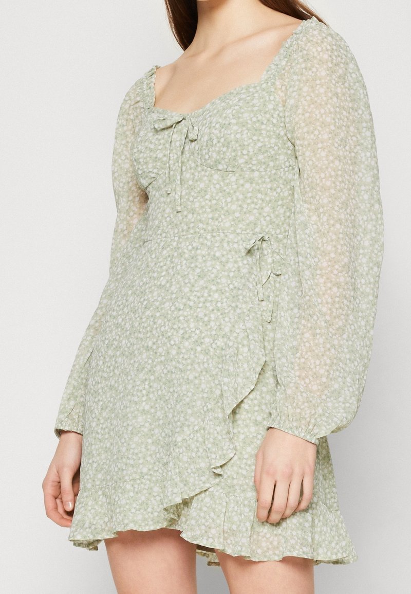 Ljusgrön blommig klänning med långa genomskinliga puffärmar, hjärtformad halsringning, frontdetaljer med knytning och volangkant i asymmetrisk stil, bärs av en person.