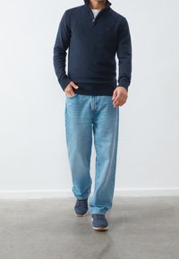 Man klädd i marinblå kvartzip-tröja, ljusblå jeans och marinblå sneakers som står med en hand i fickan på en grå golv.