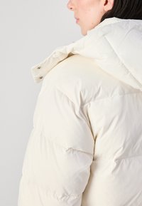 Hvit pufferjakke med quiltet design, hette og synlige detaljer. Materialet ser glatt og vannavstøtende ut.