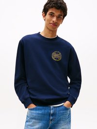 Felpa di cotone blu navy con scollo rotondo e polsini a costine, caratterizzata da un logo ricamato in oro sul petto e abbinata a jeans azzurri.