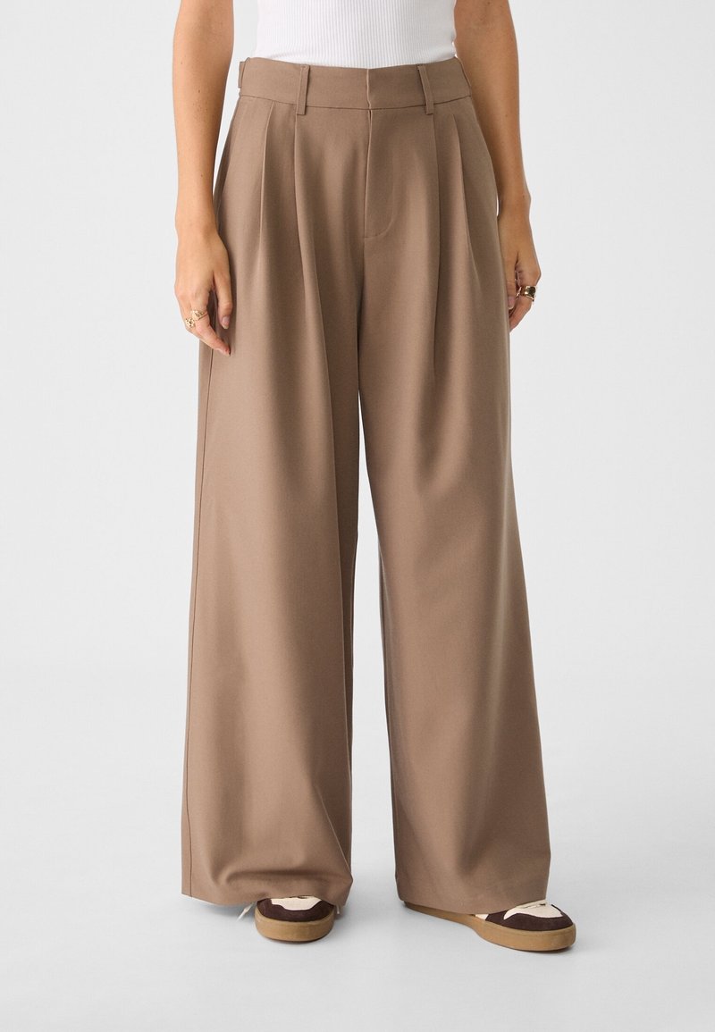 Femme portant un pantalon marron taille haute à jambes larges, associé à des baskets blanches et marron, debout devant un fond clair uni.