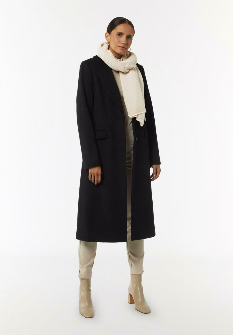 Czarny długi płaszcz z klapami, beżowy tkany szalik, kremowe spodnie oraz jasne, wysokie buty na obcasie. Gładkie tekstury i dopasowany krój.